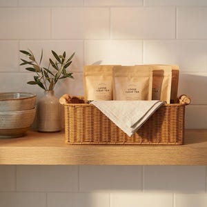 Pode incluir: Uma cesta retangular de vime com saquinhos de chá de papel castanho e um pano de linho dobrado. A cesta está numa prateleira de madeira, com um vaso de vegetação e tigelas de cerâmica à esquerda. Os saquinhos de chá estão rotulados como "Loose Leaf Tea."