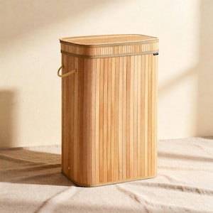 May include: A rectangular wooden laundry hamper with a lid. The hamper is made from vertical wooden slats, with a light brown colour. It has a matching wooden lid and a tan fabric trim. A tan handle is on the side.