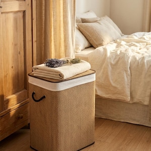 May include: A rectangular woven laundry hamper with a dark brown trim and handle. A folded beige blanket and a bundle of lavender rest on top. The hamper is in a bedroom setting with a bed and wooden furniture.