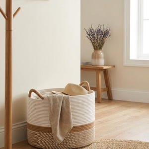Può includere: Un cesto rotondo intrecciato con un design a corda bianca e beige. Il cesto ha due manici e contiene un asciugamano beige e un cappello di paglia. Un piccolo tappeto rotondo è sul pavimento davanti al cesto. Un attaccapanni in legno è a sinistra.