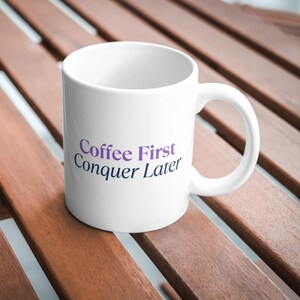 以下が含まれることがあります： 紫と青で「Coffee First Conquer Later」と書かれた白いセラミックコーヒーマグ。 マグカップには湾曲したハンドルがあり、木製の表面に置かれています。