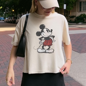 K&ouml;nnte beinhalten: Cremefarbenes T-Shirt mit einem Mickey Mouse-Motiv in Schwarz, Rot und Wei&szlig;. Das Shirt hat kurze &Auml;rmel und eine lockere Passform. Das Wort "IVORY" ist unten links im Bild aufgedruckt. Die Person tr&auml;gt eine beige Kappe und einen schwarzen Rock.