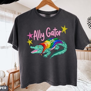 May include: Dark gray t-shirt with the text "Ally Gator" in pink and yellow stars. The shirt features a cartoon alligator with a rainbow Pride flag. The alligator is teal and green.