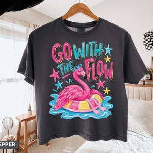 May include: A charcoal gray t-shirt featuring a graphic of a pink flamingo wearing blue heart-shaped sunglasses while floating on a yellow and pink inner tube in blue water, with the text "GO WITH THE FLOW" and colorful stars surrounding it.