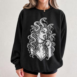 Puede incluir: Una sudadera negra de manga larga con un gran estampado gráfico en blanco de una figura con serpientes por cabello, que lleva gafas de sol y un collar con calavera, mientras sostiene una piruleta. El diseño está centrado en el pecho.