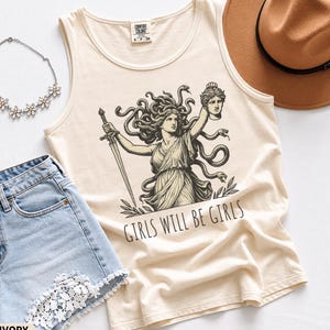 Könnte beinhalten: Ein elfenbeinfarbenes Tanktop mit einer schwarzen grafischen Illustration von Medusa, die ein Schwert und einen abgetrennten Kopf hält, mit dem Text "GIRLS WILL BE GIRLS" darunter. Das Kleidungsstück hat einen Rundhalsausschnitt und ein Comfort Colors Etikett.