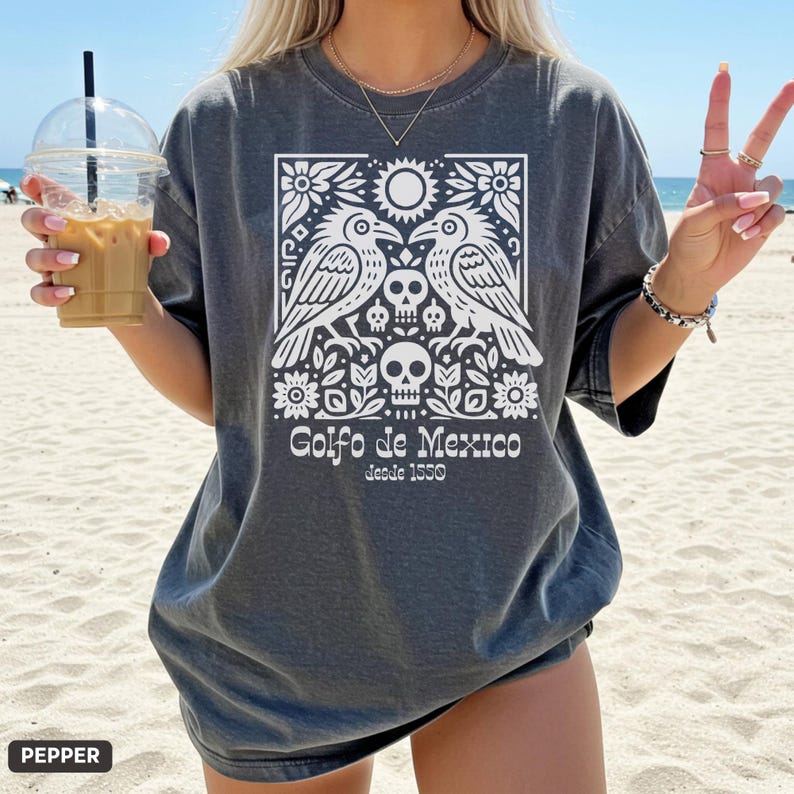 Puede incluir: Camiseta gris oscuro con un dise&ntilde;o gr&aacute;fico blanco que muestra dos aves, calaveras, flores y el texto "Golfo de Mexico desde 1550". La camiseta es de corte holgado y tiene mangas cortas.