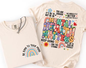 Könnte beinhalten: Zwei elfenbeinfarbene T-Shirts. Eines zeigt einen Regenbogen und den Text "BE KIND TO YOUR MIND". Das andere zeigt die Worte "MENTAL HEALTH MATTERS" in einem farbenfrohen, floralen Design mit Sätzen wie "KEEP GOING" und "END THE STIGMA."