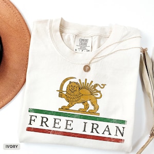 Puede incluir: Camiseta de color marfil con un dise&ntilde;o de le&oacute;n y sol, las palabras "FREE IRAN" en rojo y verde, y un collar dorado. La camiseta est&aacute; doblada y se ve parcialmente un sombrero marr&oacute;n.