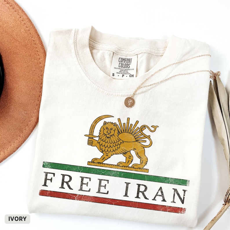 Puede incluir: Camiseta de color marfil con un dise&ntilde;o de le&oacute;n y sol, las palabras "FREE IRAN" en rojo y verde, y un collar dorado. La camiseta est&aacute; doblada y se ve parcialmente un sombrero marr&oacute;n.