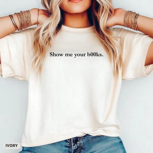 以下が含まれることがあります： 「Show me your books.」の黒い文字が入ったアイボリー色のTシャツ。クルーネックと半袖です。Tシャツの左下に「IVORY」の文字がプリントされています。