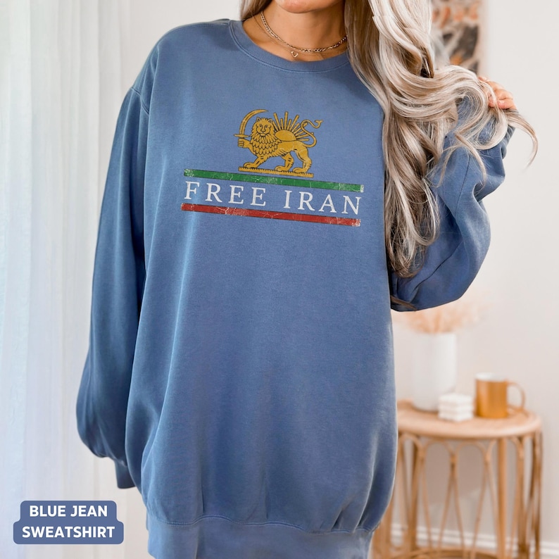 Puede incluir: Sudadera vaquera azul con el texto blanco "FREE IRAN" sobre una franja verde y roja. La sudadera presenta un emblema dorado de un le&oacute;n con un sol.