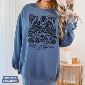 Puede incluir: Sudadera azul vaquera con un dise&ntilde;o gr&aacute;fico negro que presenta dos p&aacute;jaros, un sol, flores y calaveras. El texto "Golfo de Mexico desde 1550" est&aacute; impreso debajo. Manga larga.