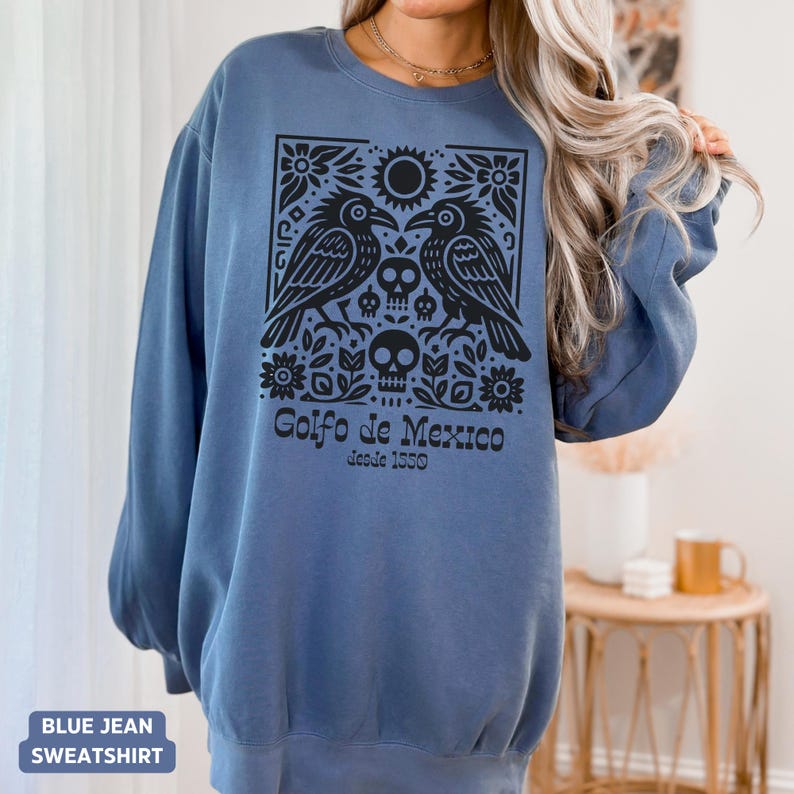 Puede incluir: Sudadera azul vaquera con un dise&ntilde;o gr&aacute;fico negro que presenta dos p&aacute;jaros, un sol, flores y calaveras. El texto "Golfo de Mexico desde 1550" est&aacute; impreso debajo. Manga larga.