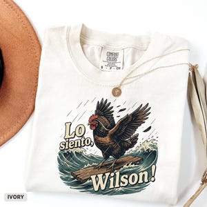 Könnte beinhalten: Elfenbeinfarbenes T-Shirt mit einem Grafikdesign eines surfenden Hahns mit dem Text "Lo siento, Wilson!". Das Design zeigt Wellen und Regen. Das T-Shirt hat einen Rundhalsausschnitt und kurze Ärmel. Eine goldene Halskette und ein brauner Hut sind ebenfalls sichtbar.
