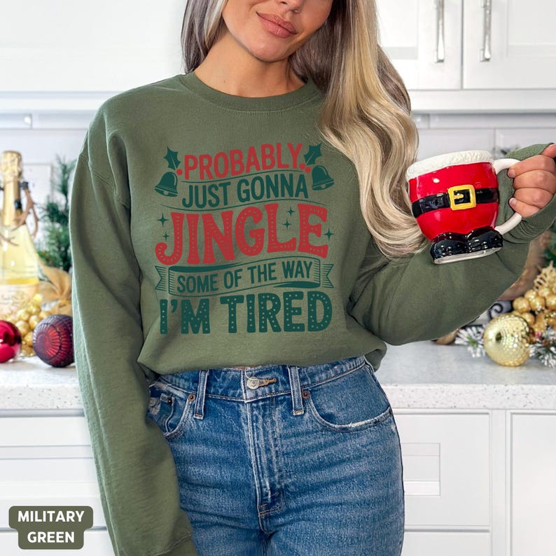 May include: Military green sweatshirt with the text "Probably Just Gonna Jingle Some of the Way I'm Tired" in red and green. The person is wearing blue jeans and holding a Santa-themed mug.