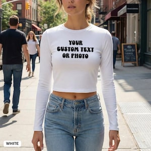 May include: A white, long-sleeved crop top with the text "YOUR CUSTOM TEXT OR PHOTO" in black. The top is worn with light blue jeans. The word "WHITE" is visible at the bottom left of the image.