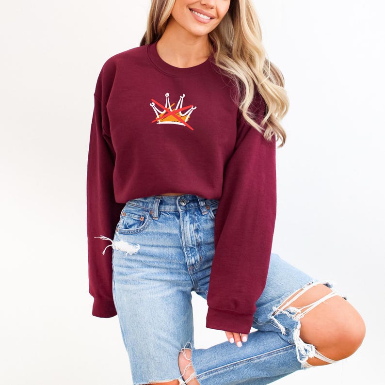 Puede incluir: Una sudadera granate con un peque&ntilde;o dise&ntilde;o de corona en el pecho. La corona tiene detalles en blanco, amarillo y rojo. La sudadera se combina con vaqueros azul claro con detalles desgastados.