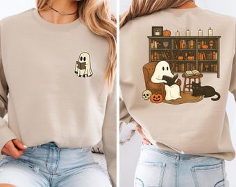 Puede incluir: Sudadera beige con un fantasma de dibujos animados leyendo un libro en la parte delantera. La parte trasera de la sudadera muestra un fantasma leyendo en una silla frente a una estantería, con un gato negro, calabazas y velas. Un diseño temático de Halloween.