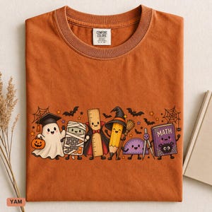May include: A burnt orange t-shirt featuring a graphic of cartoon school supplies dressed for Halloween, including a ghost with a pumpkin, a mummy calculator, a vampire ruler, a witch pencil, a protractor, and a book labeled MATH.