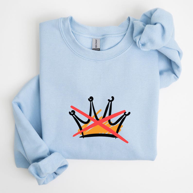 Puede incluir: Sudadera azul claro con un dise&ntilde;o de corona dibujada a mano con una 'X' roja. La corona est&aacute; delineada en negro con un centro naranja. La sudadera est&aacute; hecha de un material suave.