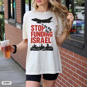 Op de afbeelding: Ivoor kleurig t-shirt met een zwarte afbeelding van een straalvliegtuig, dollartekens en de rode tekst "STOP FUNDING ISRAEL". Onder de tekst staat een zwart silhouet van een stadsgezicht. Het shirt wordt gedragen.