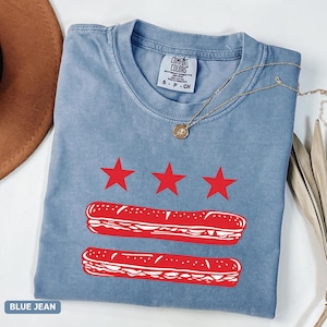 May include: A blue jean colored t-shirt with a red graphic design. The design includes three red stars above two red submarine sandwich illustrations. The shirt has a crew neck and short sleeves. The brand name "Comfort Colors" is visible on the shirt.