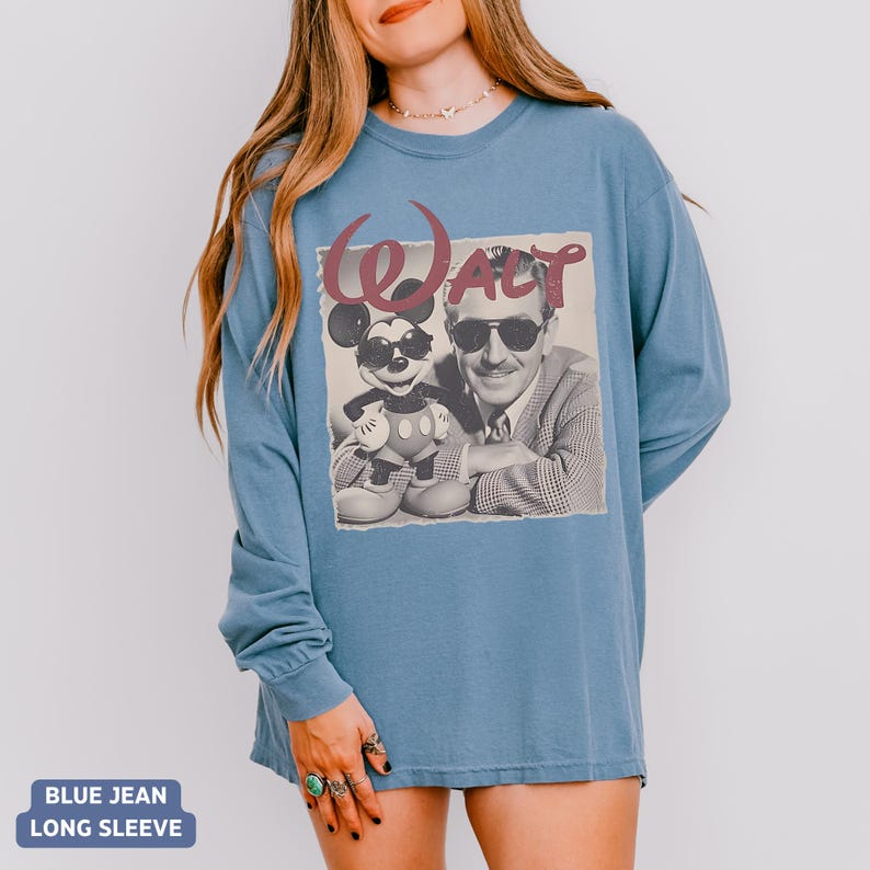May include: A blue long-sleeve t-shirt featuring a graphic print of Walt Disney and Mickey Mouse both wearing sunglasses. The word Walt is printed in a large, stylized maroon font above the black and white image of the two figures.