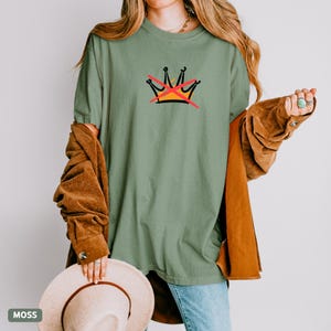 May include: A moss green t-shirt featuring a graphic of a crown with a red X through it. The shirt is a relaxed fit and has short sleeves. The word "MOSS" is in a small box in the lower left corner.