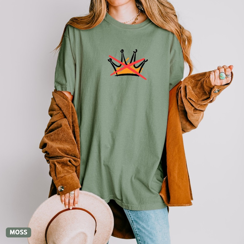 May include: A moss green t-shirt featuring a graphic of a crown with a red X through it. The shirt is a relaxed fit and has short sleeves. The word "MOSS" is in a small box in the lower left corner.