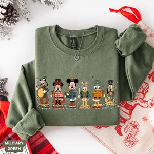 May include: Military green sweatshirt featuring a line of Disney characters dressed as toy soldiers. The characters include Mickey Mouse, Minnie Mouse, Donald Duck, and Goofy. The design is festive and holiday-themed.