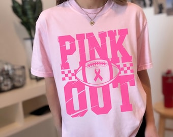 Puede incluir: Una camiseta rosa claro con el texto PINK OUT en letras fucsias, negritas y desgastadas. En el centro, un gráfico de un balón de fútbol americano con un lazo rosa de concienciación, flanqueado por pequeños cuadros a ambos lados.