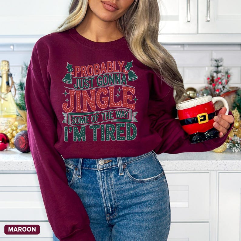 May include: Maroon sweatshirt with the text "PROBABLY JUST GONNA JINGLE SOME OF THE WAY I'M TIRED" in green and red. The person is holding a Santa-themed mug. The sweatshirt is paired with blue jeans.
