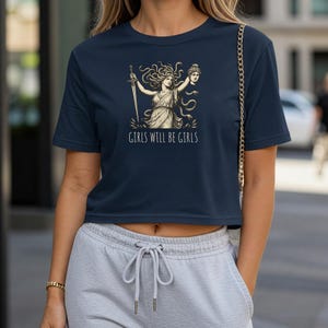 May include: Navy blue crop top featuring a graphic of a woman with a sword and a severed head, with the text "GIRLS WILL BE GIRLS" in white. The design is in a vintage style. The top is a casual, short-sleeved shirt.