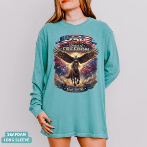 May include: Seafoam green long-sleeve shirt featuring a graphic of an eagle with outstretched wings, a horse, and the text "250 Years of Freedom Est. 1776" in a patriotic design. The shirt is a comfortable, casual style.