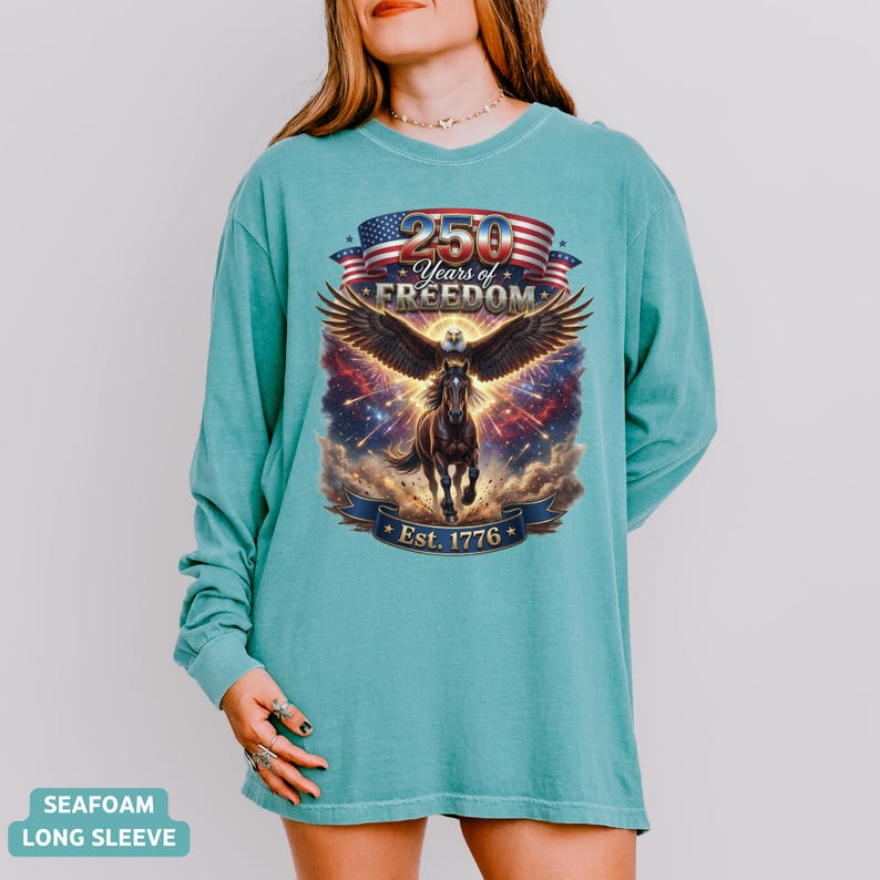 May include: Seafoam green long-sleeve shirt featuring a graphic of an eagle with outstretched wings, a horse, and the text "250 Years of Freedom Est. 1776" in a patriotic design. The shirt is a comfortable, casual style.