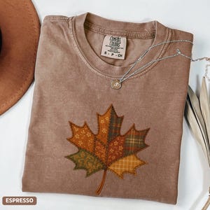 May include: Espresso-coloured t-shirt with a patchwork maple leaf design. The leaf features various patterns in shades of orange, green, and brown. A silver necklace with a pendant rests on the shirt. The shirt is labelled "Comfort Colors".