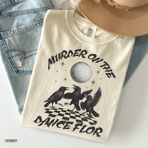 May include: Ivory-colored t-shirt with the text "MURDER ON THE DANCE FLOR" and a graphic of three crows, a disco ball, and stars. The shirt is made of a soft material and is a Comfort Colors brand.