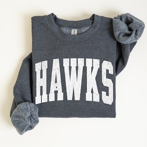 May include: A dark gray sweatshirt with the word "HAWKS" printed in large, distressed white letters across the front. The sweatshirt has a crew neck and long sleeves, with the sleeves cuffed. The fabric appears soft and comfortable.