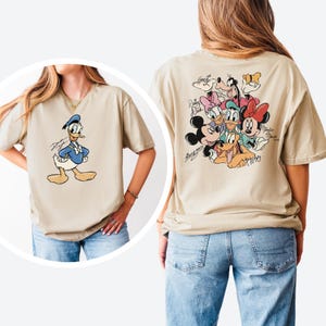 Pode incluir: Camiseta bege com o Pato Donald na frente e um grupo de personagens da Disney nas costas. O Pato Donald &eacute; retratado em um traje de marinheiro azul. A parte de tr&aacute;s da camisa apresenta Mickey Mouse, Minnie Mouse, Pateta, Pluto, Margarida e Pato Donald.