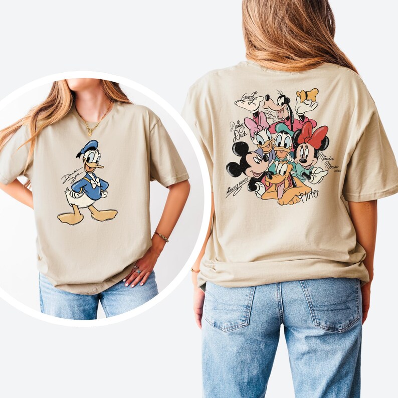 Pode incluir: Camiseta bege com o Pato Donald na frente e um grupo de personagens da Disney nas costas. O Pato Donald &eacute; retratado em um traje de marinheiro azul. A parte de tr&aacute;s da camisa apresenta Mickey Mouse, Minnie Mouse, Pateta, Pluto, Margarida e Pato Donald.