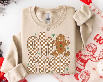 Könnte beinhalten: Ein sandfarbenes Sweatshirt mit dem Schriftzug "Oh Snap" in einem braun-weißen Schachbrettmuster und einer Lebkuchenmann-Grafik. Das Design enthält grüne und rote Akzente, ideal für die Weihnachtszeit. Das Gildan-Markenschild ist sichtbar.