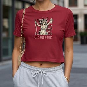 May include: A maroon crop top featuring a graphic of a woman with snakes for hair, holding a sword and a severed head. The text "GIRLS WILL BE GIRLS" is printed below the graphic. The top is paired with gray sweatpants.