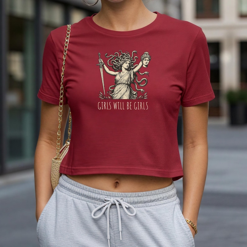 May include: A maroon crop top featuring a graphic of a woman with snakes for hair, holding a sword and a severed head. The text "GIRLS WILL BE GIRLS" is printed below the graphic. The top is paired with gray sweatpants.