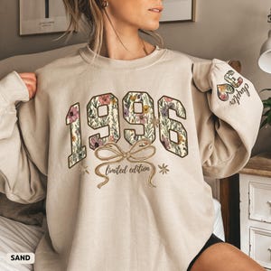 May include: Sand-colored sweatshirt with the year "1996" in a floral design, a bow, and the words "limited edition." The sleeve features the text "36 chapter" with floral accents. The word "SAND" is at the bottom left.