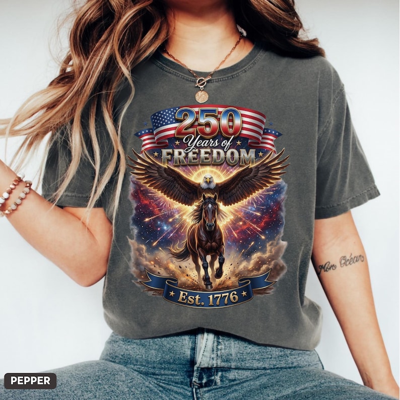 May include: Dark gray t-shirt featuring a patriotic design. The graphic includes an eagle with outstretched wings, a horse, and the text "250 Years of Freedom Est. 1776" with an American flag motif. The shirt is a casual wear item.