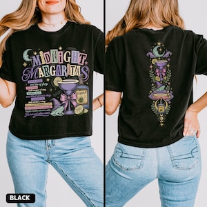May include: A black t-shirt featuring a graphic print on the front and back. The front displays the text Midnight Margaritas with a margarita glass, a frog wearing a witch hat, and a tequila bottle. The back shows a vertical design of a crescent moon, a margarita glass, a frog on books, and a tequila bottle.