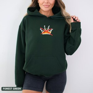 Puede incluir: Sudadera con capucha verde bosque con bolsillo delantero y capucha con cord&oacute;n. La sudadera tiene un peque&ntilde;o dise&ntilde;o de corona bordada con una l&iacute;nea roja. El texto "FOREST GREEN" est&aacute; en la parte inferior de la imagen.