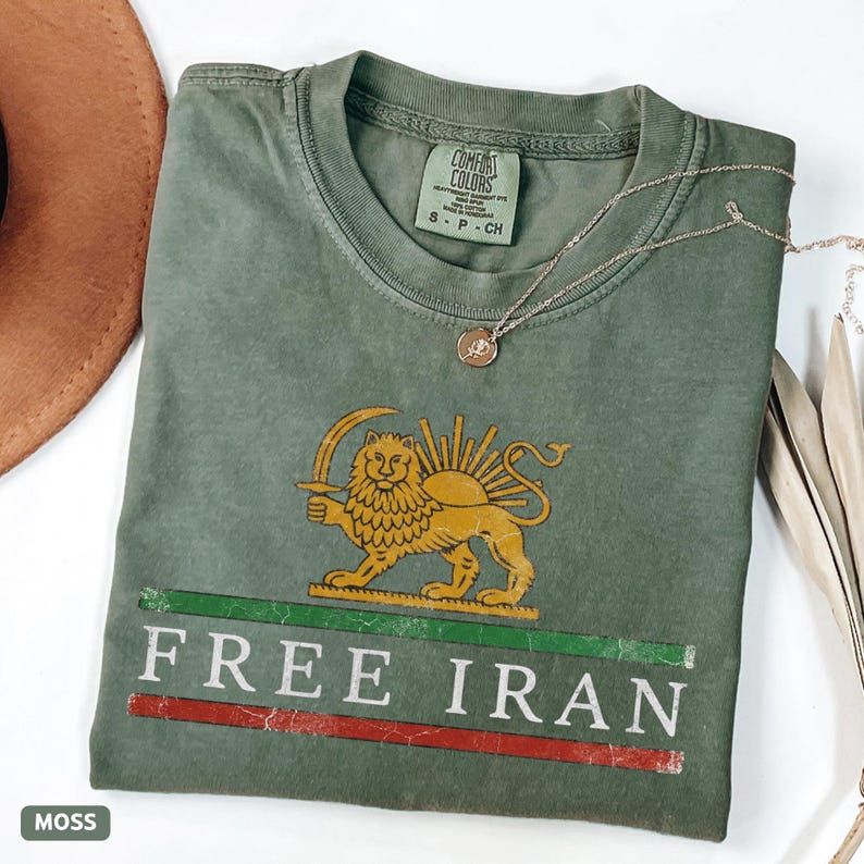 Puede incluir: Camiseta verde musgo con un le&oacute;n dorado y un gr&aacute;fico de sol, un collar plateado y el texto "FREE IRAN" en blanco. La camiseta tiene un aspecto desgastado y una peque&ntilde;a etiqueta "Comfort Colors".