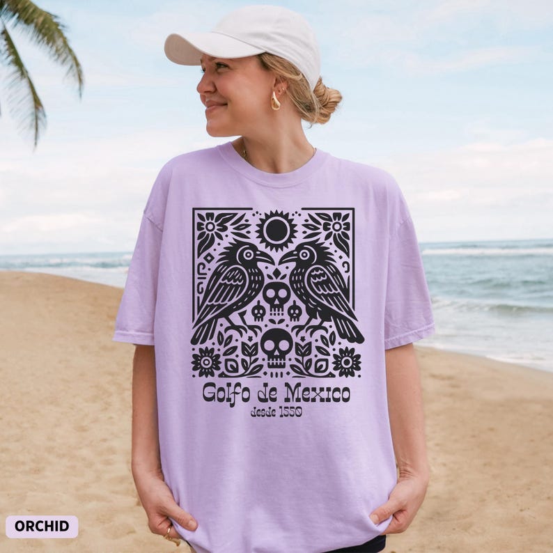 Puede incluir: Camiseta lila claro con un dise&ntilde;o gr&aacute;fico negro de dos p&aacute;jaros, calaveras y elementos florales. El texto "Golfo de Mexico desde 1550" est&aacute; impreso debajo. La camiseta est&aacute; etiquetada como "ORCHID".